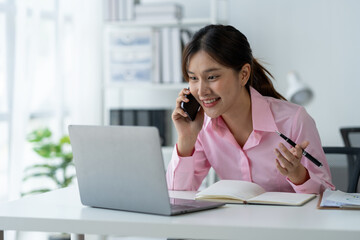 Happy young asian woman talking on the mobile phone and smiling while sitting at her working place in office.