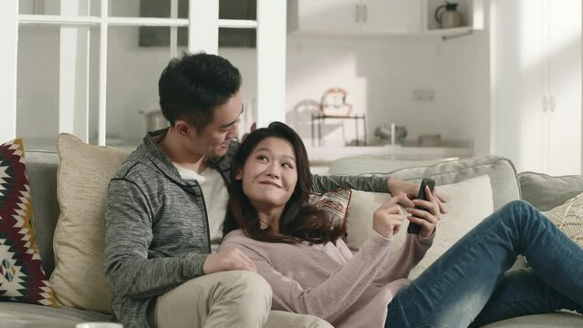young asian couple sitting lying on couch at home looking at cellphone having a pleasant conversation