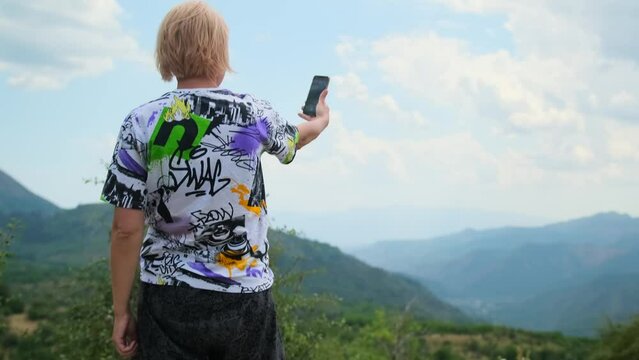 Inspired young woman tourist takes photo smartphone selfie against backdrop of breathtaking mountain landscape on top mountain in tourist travel. 