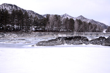 Stone formations in the middle of the frozen bed of a beautiful river flowing through a snowy mountain valley on a cloudy winter morning.