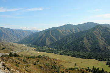 Obraz premium A winding road descends into a picturesque valley surrounded by mountains overgrown with coniferous forests on a clear summer day.