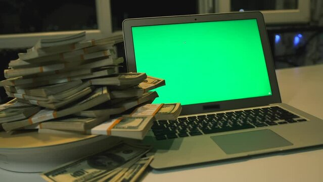 100 American dollar bills big pyramid on rotating surface next to laptop green screen on desk in office night time. Cybercrime concept. Exchange currency market.