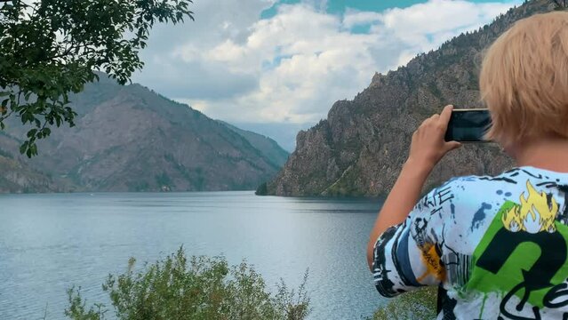 Tourist woman blonde shoots on phone scenic views clear lake and mountains, make video grandiose landscape in tourist trip for your blog. Relax and travel concept.