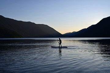 Male paddleboarder in silhouette at sunset on Lake Crescent in Olympic National Park, Washington on calm clear summer afternoon.