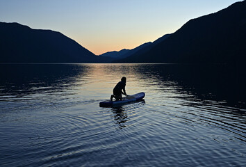 Female paddleboarder in silhouette at sunset on Lake Crescent in Olympic National Park, Washington on calm clear summer afternoon.