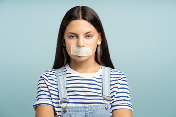 Unhappy teenage girl with closed mouth sealed white tape isolated on studio blue background. Sad...
