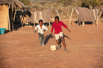 village african kids playing soccer on an improvised football field in the yard between the thatched huts homes - obrazy, fototapety, plakaty