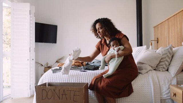 Black Woman Holds Baby while Sorting Clothes for Donation Indoors