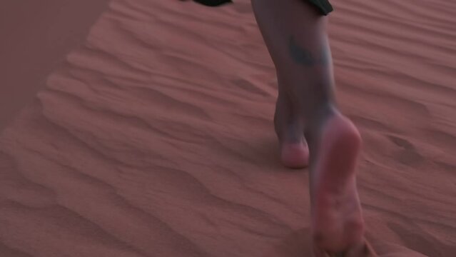 A swarthy young woman in a long black dress and an Arab scarf walks barefoot on the desert sand to a white SUV standing on a dune