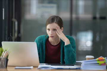 A tired businesswoman under stress works at a laptop while sitting at a table at home and holds her hand on her temples, causing migraine attacks.