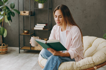 Young smart happy calm woman wear casual clothes sits in armchair read book drink coffee tea stay at home hotel flat rest relax spend free spare time in living room indoor. Lifestyle lounge concept.