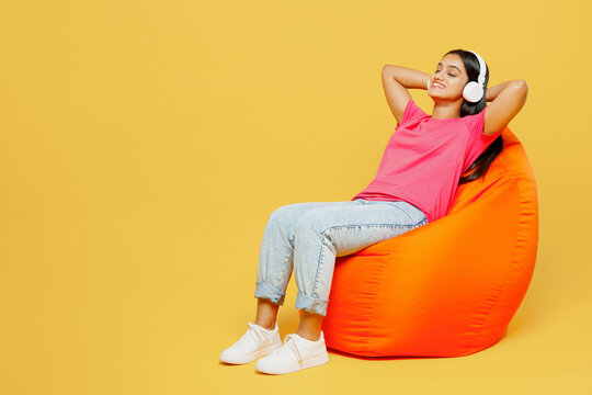 Full Body Young Happy Indian Woman Wearing Pink T-shirt Casual Clothes Sit In Bag Chair Listen To Music In Headphones Close Eyes Isolated On Plain Yellow Background Studio Portrait. Lifestyle Concept.