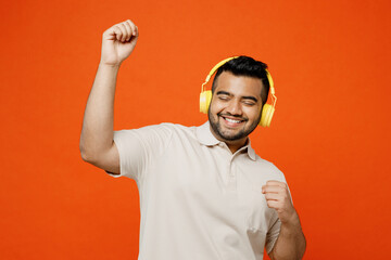 Young smiling happy fun Indian man he wears white t-shirt casual clothes listen to music in headphones raise up hands dance isolated on plain orange red background studio portrait. Lifestyle concept.