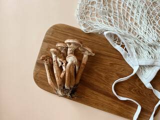 Mushrooms of honey mushrooms lie on a wooden oak board on a beige background next to a white braided string bag. Mushroom picking. Copy space. Top view