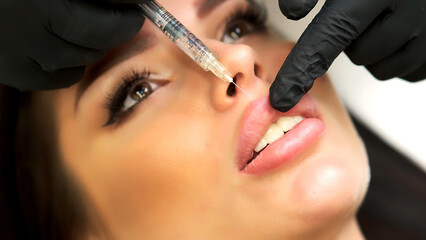 A beautician in medical gloves carefully and slowly injects hyaluronic acid into the lips of a...