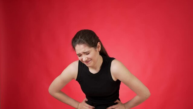 Distressfully looking casually dressed young woman with long dark hair touching her tummy with both hands apparently experiencing acute stomach pain against red background