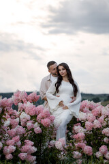 Pregnant young romantic couple in love standing among flowers in field. An attractive boy and a tender girl are hugging and holding a pregnant belly in their hands, the feeling of happiness is present