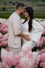 Pregnant young romantic couple in love standing among flowers in field. Attractive boy and tender girl hug, look at each other and hold pregnant belly in hands, present feeling of happiness. Love, fam