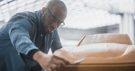 Portrait of a Handsome Automotive Designer Developing a 3D Clay Model of a New Production Car. Professional Black Man Using a Spatula to Smooth Out and Trim the Surface of a Prototype Concept Car