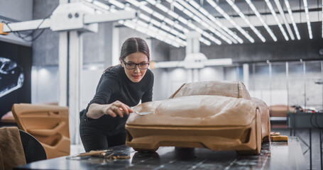 Portrait of a Female Automotive Designer Sculpting a 3D Clay Model of New Production Car. Young Woman Using a Spatula to Carefully Trim the Surface of a Prototype Concept Vehicle