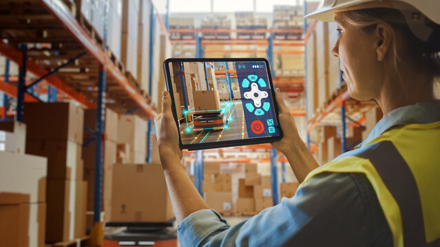Future Technology Concept: Female Worker Holding Tablet With AR Remote Control Application For Retail Warehouse AGV Robot with Infographics. Women Controlling Automated Guided Vehicles With Products. - Powered by Adobe
