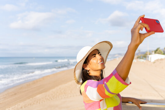 Beautiful latina woman with a hat and flashy and colorful dress enjoys a beautiful summer guide from the top of a lifeguard watchtower doing a selfie.