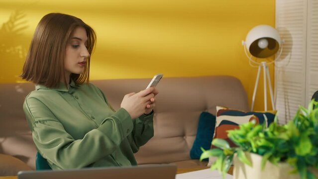 Side view of focused girl texting message on smartphone at home. Thoughtful woman browsing internet on cellphone. Serious lady chatting on mobile phone. Smart student relaxing after online lessons