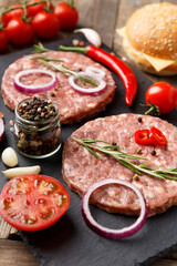 Raw beef hamburger patties on stone plate, vegetables and spices, wooden background.