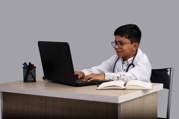 Portrait Indian Asian kid boy aged 7 to 8, wearing a doctor apron with stethoscope. with laptop writing prescription. He had a dream to future study as Doctor. He smiled happily, Concept little Doctor