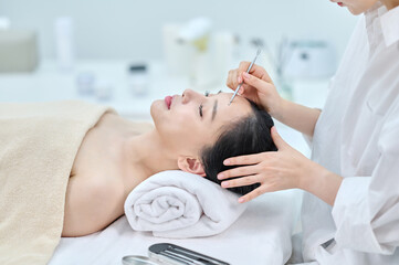 아시아 한국의 젊은 여성이 스파 또는 피부관리전문점 또는 피부과 병원에서 침대에 누워 베이지색 대형 타올을 덮고 눈을 감고있고 하얀색 셔츠를 입은 전문관리사가 여드름 압출기로 문제성피부를 관리하고 있다. 