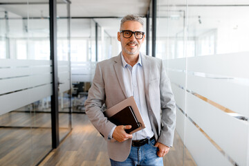 Portrait of charismatic senior middle aged man in formal wear carrying laptop in office hallway,...