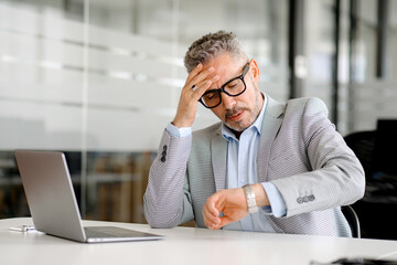 Disappointed mature businessman in formal wear looking at the watch on his wrist and worrying about...