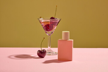 Front view of pink mockup bottle unbranded decorated with a glass cup and fresh ripe cherries on color background. Scene for advertising product with cherry extracted. Space for design