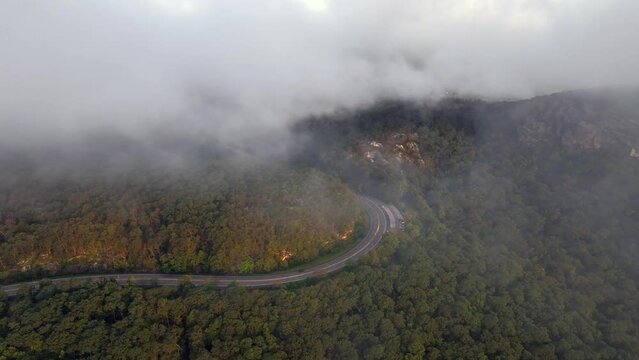 A beautiful aerial shot over Storm King Mountain in Upstate New York. Taken after sunrise with low lying clouds. The drone camera dolly in and boom down through the clouds towards an empty Route 9W.