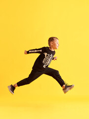 Fototapeta na wymiar Full-length image of little playful boy, child in black costume jumping against yellow studio background. Game. Concept of childhood, kids emotions, Halloween party, joy and fun, fashion, ad