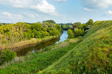 Obraz premium View from a hill with steep slopes to the river with deciduous trees on the banks on a sunny summer day. Landscape of beautiful and picturesque nature