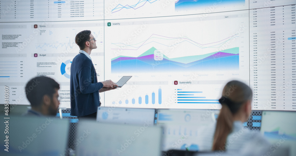 Aufkleber Male Senior Data Analyst Reviewing Financial Information And Reports on Big Digital Screen in Monitoring Office. Diverse Employees Working Behind Desktop Computers In Venture Capital Company.