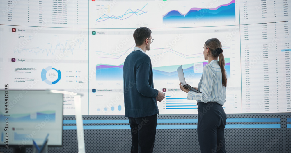 Poster Back View Following Shot Of Male And Female Analysts Discussing Data On Big Digital Screen In Monitoring Office. Diverse Employees Working Behind Desktop Computers In Artificial Intelligence Company.