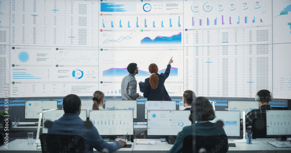 Poster Back View Of Indian Data Analyst And Hispanic Manager Discussing Reports On Digital Screen in Monitoring Room. Male and Female Experts Talking, Employees Working On Computers In Consulting Company