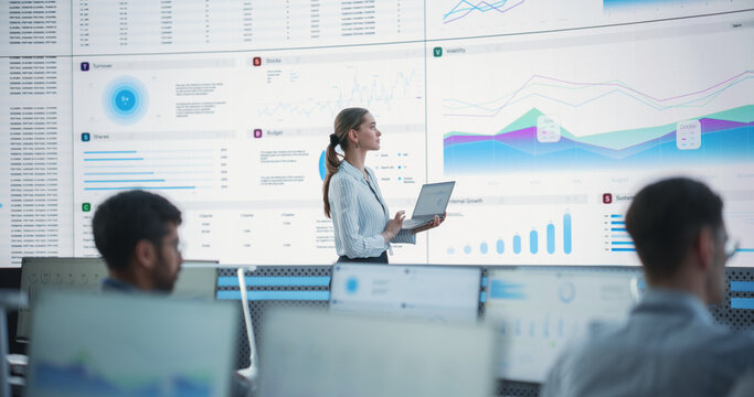 Female Data Scientist Using Laptop And Analyzing Information And Graphs On Big Digital Screen In Monitoring Office. Caucasian Woman Working For Big Data Company, Diverse Employees Behind Computers.