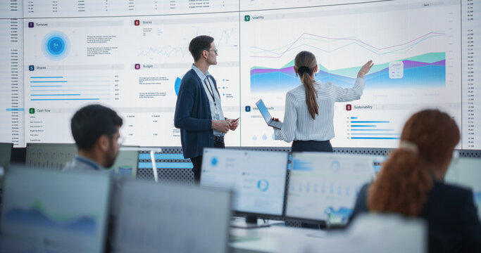 Female Data Scientist And Male Project Manager Discussing Reports And Graphs On Big Digital Screen In Monitoring Office. Diverse Employees Working Behind Computers in Artificial Intelligence Company.