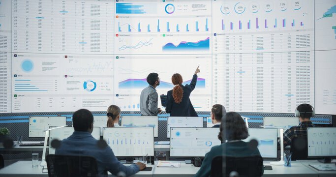 Back View Of Indian Data Analyst And Hispanic Manager Discussing Reports On Digital Screen in Monitoring Room. Male and Female Experts Talking, Employees Working On Computers In Consulting Company