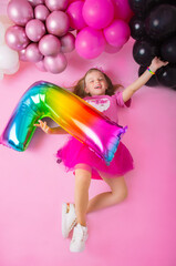 Joyful little girl lies on a pink background with balloons and holding the number 7. Happy Birthday. Pink background.