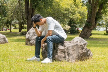 Portrait of an Asian man in a white t-shirt sitting on a rock, stressed out by problems going...