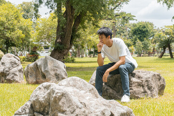 Portrait of an Asian man in a white t-shirt sitting on a rock, stressed out by problems going through life. tension concept