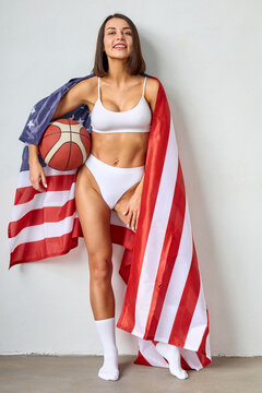 Combining athleticism and patriotism, a studio photograph captures a young woman wearing white underwear, holding a basketball, and standing beside an American flag.