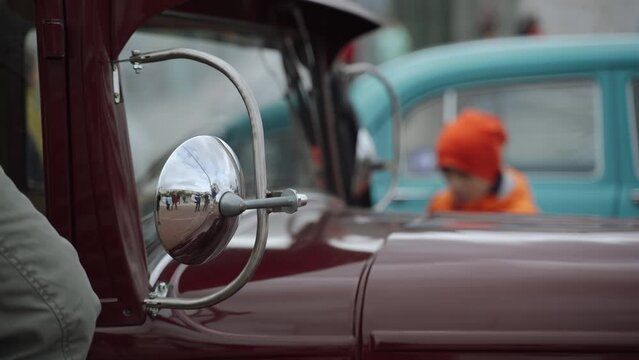 Windshield with chrome mirrors of ancient vintage car in parking lot with people. Round chrome elements of retro car on auction floor, unrecognizable people and families in warm clothes examine lot.