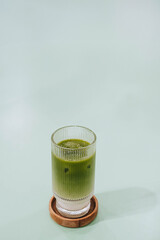 Portrait or vertical and selective focus shot of a glass of Ice Matcha or Japanese Green Tea Latte on wooden coaster on isolated sage green pastel table