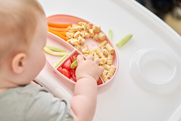 Cute child eats healthy food pasta and vegetables steamed,. Portraits of a cute 10 months old baby girl. The baby sitting in a special high chair for babies.  - 635824068