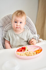 Cute child eats healthy food pasta and vegetables steamed,. Portraits of a cute 10 months old baby girl. The baby sitting in a special high chair for babies. 